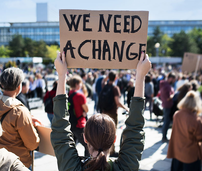 Woman at protest holds sign that reads “We Need Change”